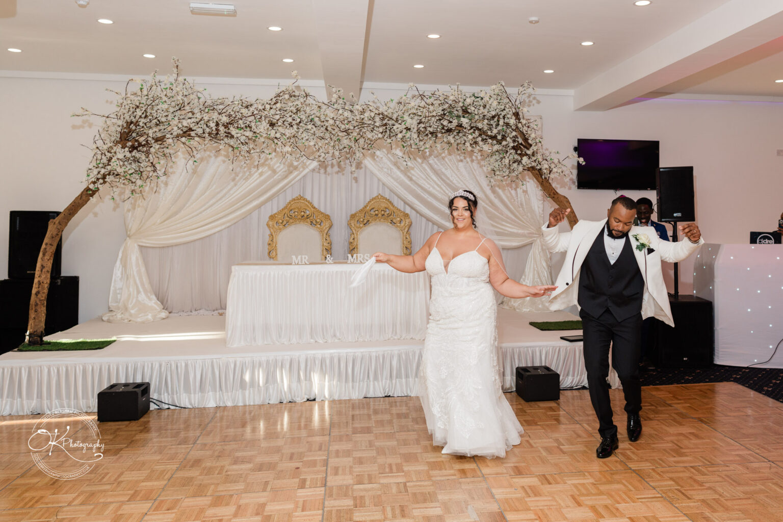 Symphony Rooms Wedding Photography Bride and groom dancing in front of a decorated stage with "Mr & Mrs" sign, white drapery, ornate chairs, and tree branch decoration overhead.