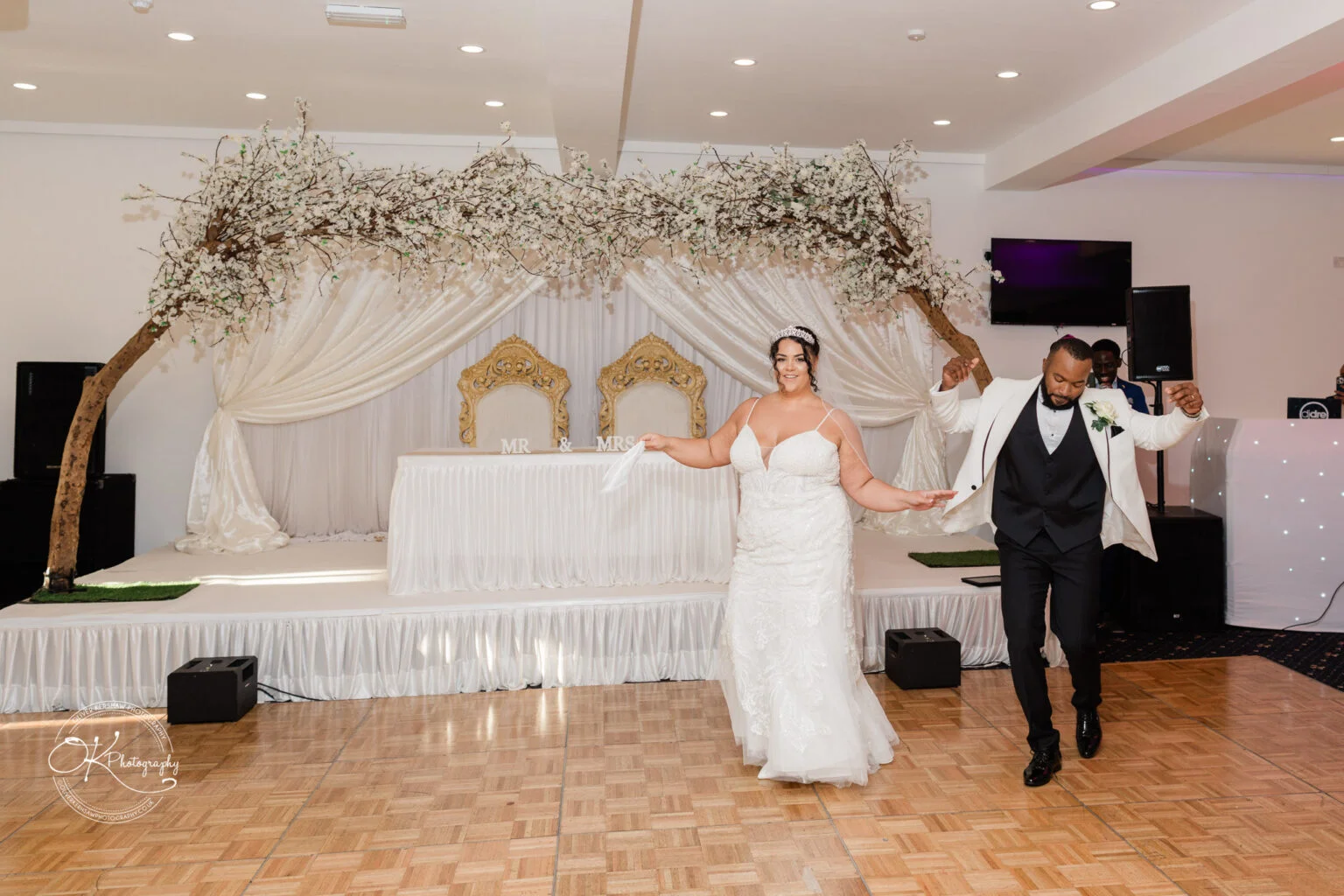 Bride and groom dancing in front of a decorated stage with "Mr & Mrs" sign, white drapery, ornate chairs, and tree branch decoration overhead.