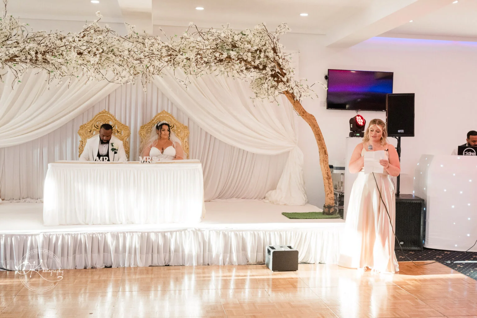 Bride and groom seated at a decorated table under a floral arch while a woman gives a speech at a wedding reception.