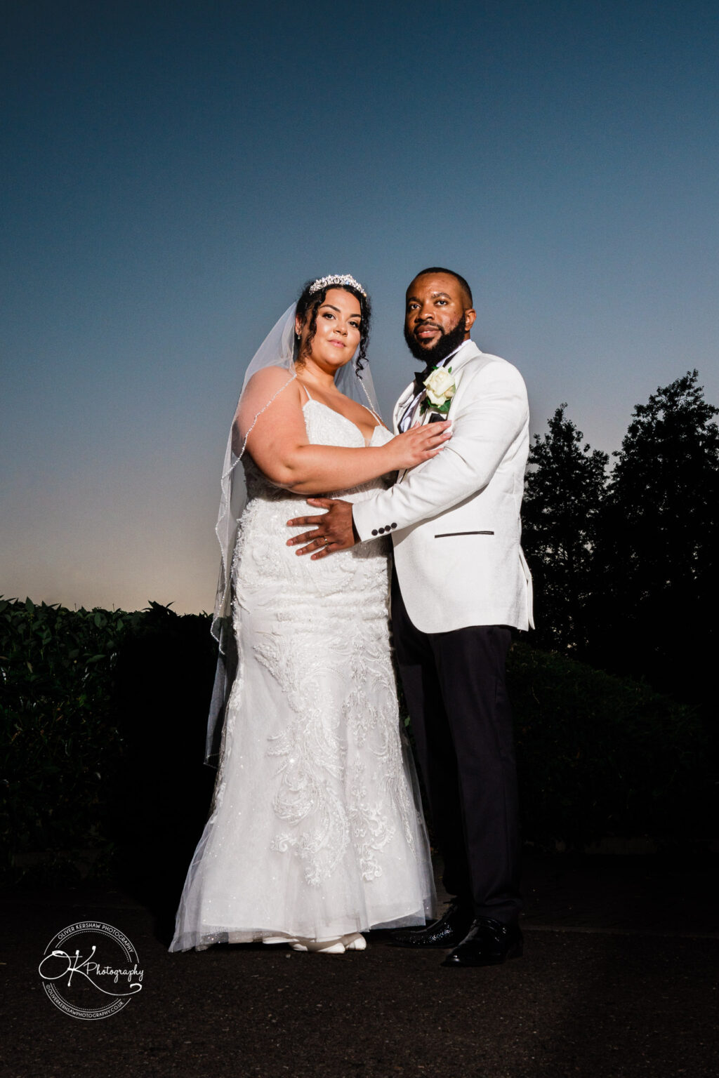 Symphony Rooms Wedding Photography Bride and groom standing together, the bride wearing a white lace wedding dress and veil, and the groom wearing a white jacket with a boutonnière, against a dusk sky backdrop with dark foliage.