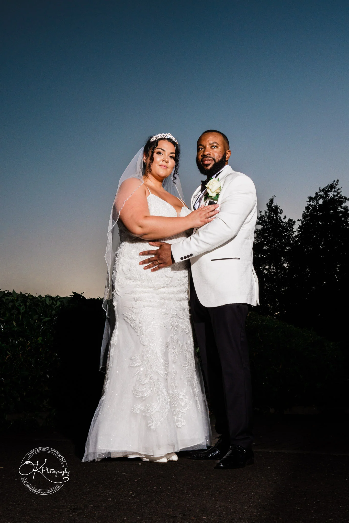 Bride and groom standing together, the bride wearing a white lace wedding dress and veil, and the groom wearing a white jacket with a boutonnière, against a dusk sky backdrop with dark foliage.