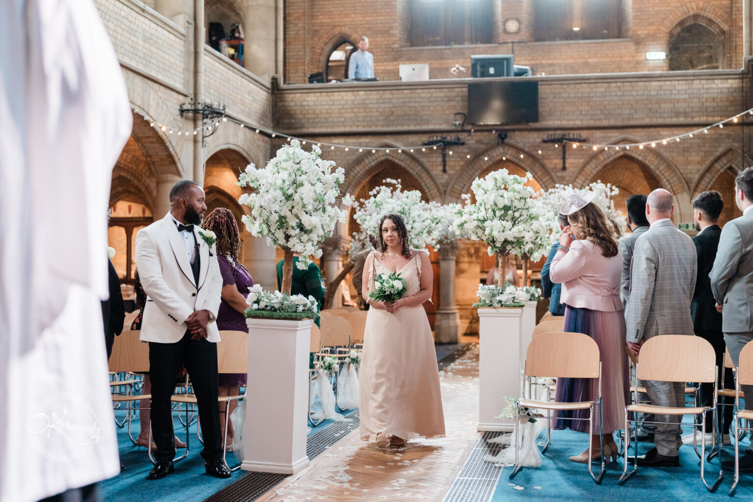 Symphony Rooms Wedding Photography A bridesmaid in a light pink dress walks down the aisle holding a bouquet, with guests seated and standing on either side, and a man in a white jacket standing at the front, in a church decorated with white floral arrangements.