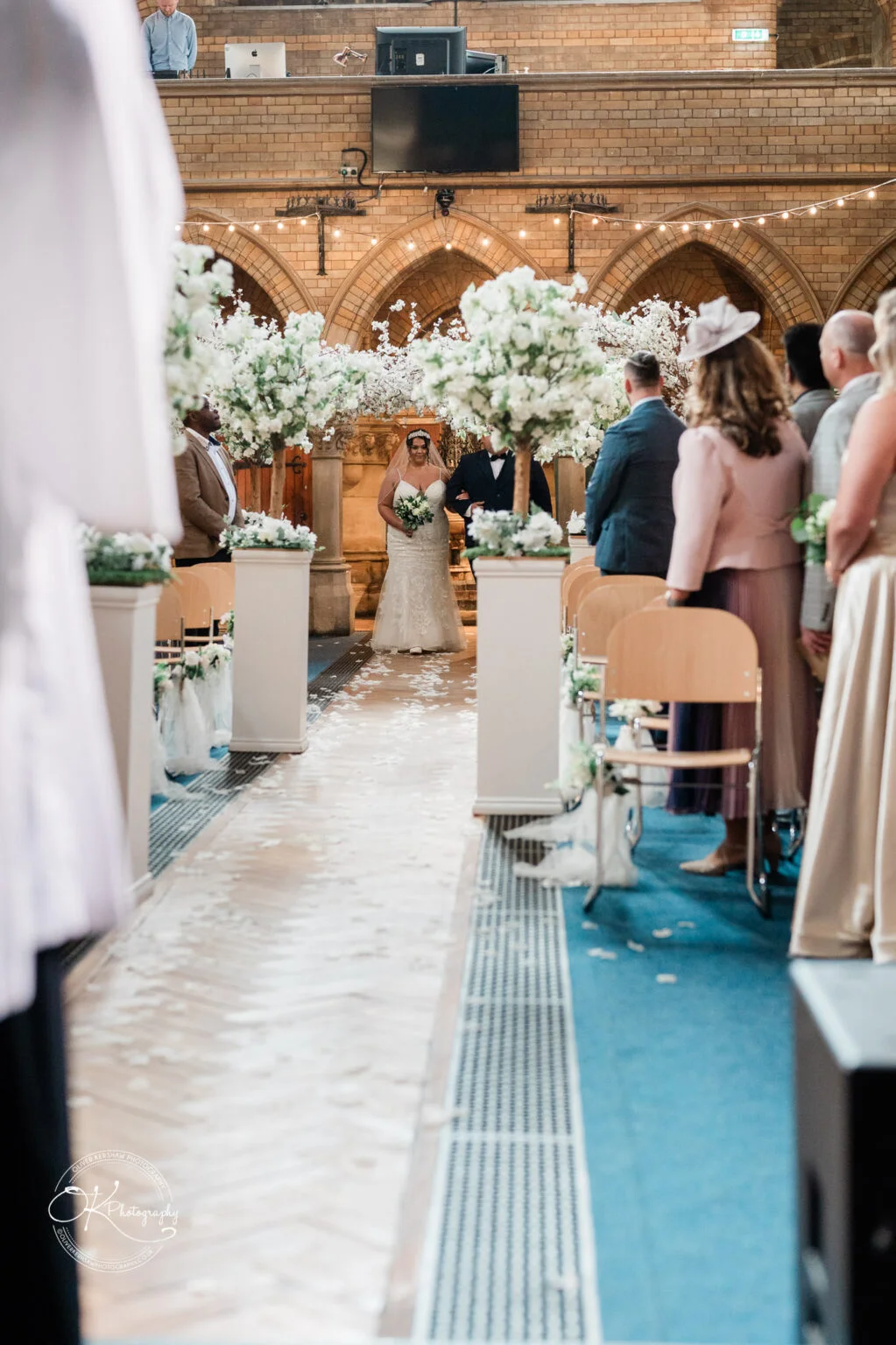 Bride walking down the aisle in a church decorated with white flowers, guests standing on either side.