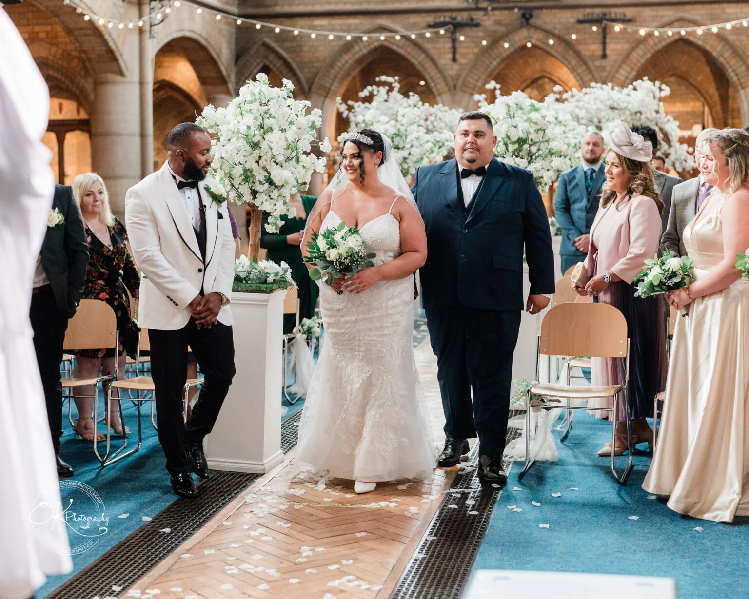 Bride in a white dress and groom in a white jacket with black bow tie standing during wedding ceremony in a church, surrounded by family and friends.