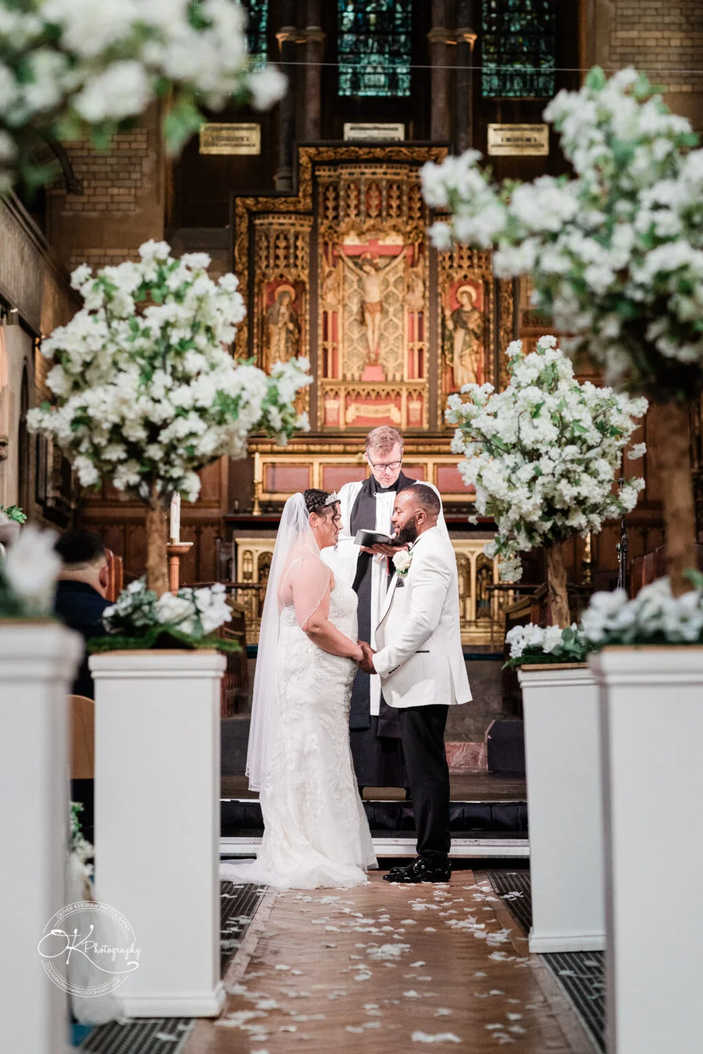 Bride and groom standing at the altar, holding hands, during their wedding ceremony in a beautifully decorated church with white flowers.