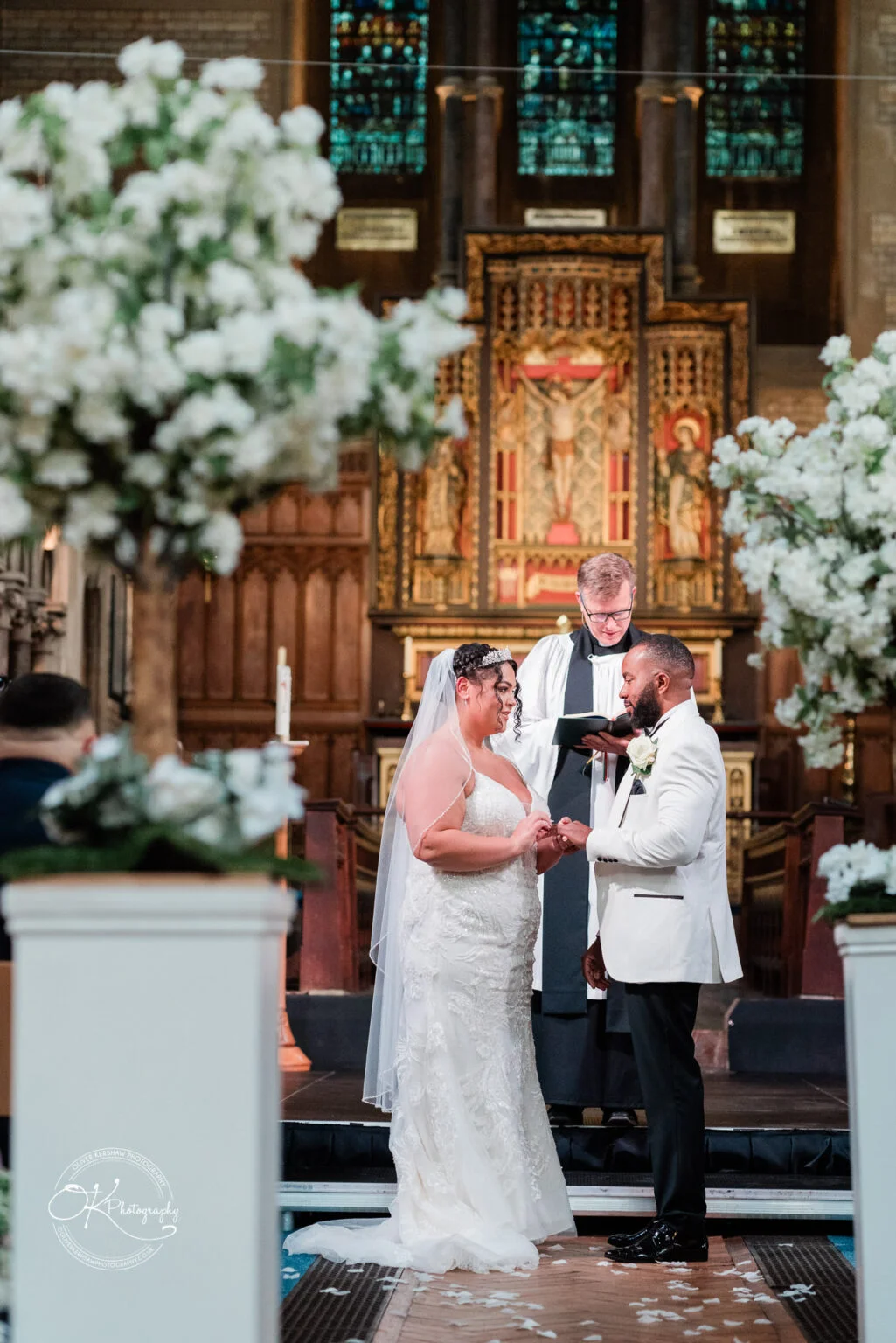 A bride and groom stand at the altar during their wedding ceremony, exchanging rings, with a priest conducting the ceremony in a church decorated with white flowers and ornate stained glass windows in the background.
