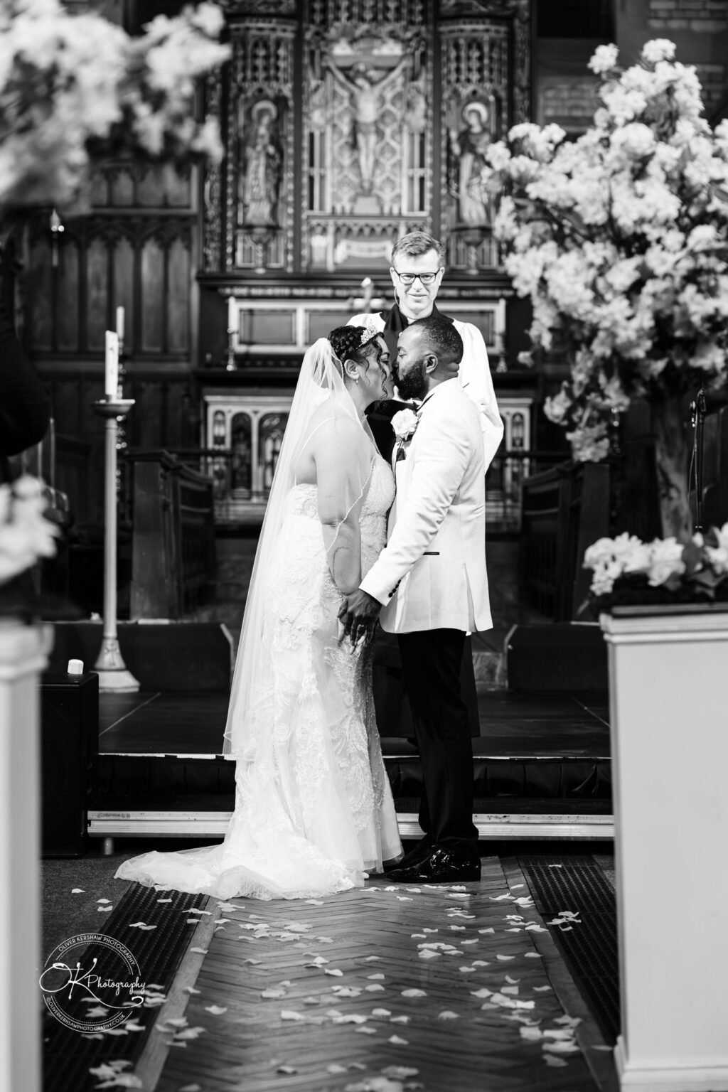 Symphony Rooms Wedding Photography A couple holding hands and kissing during their wedding ceremony in a church, with a priest standing behind them.