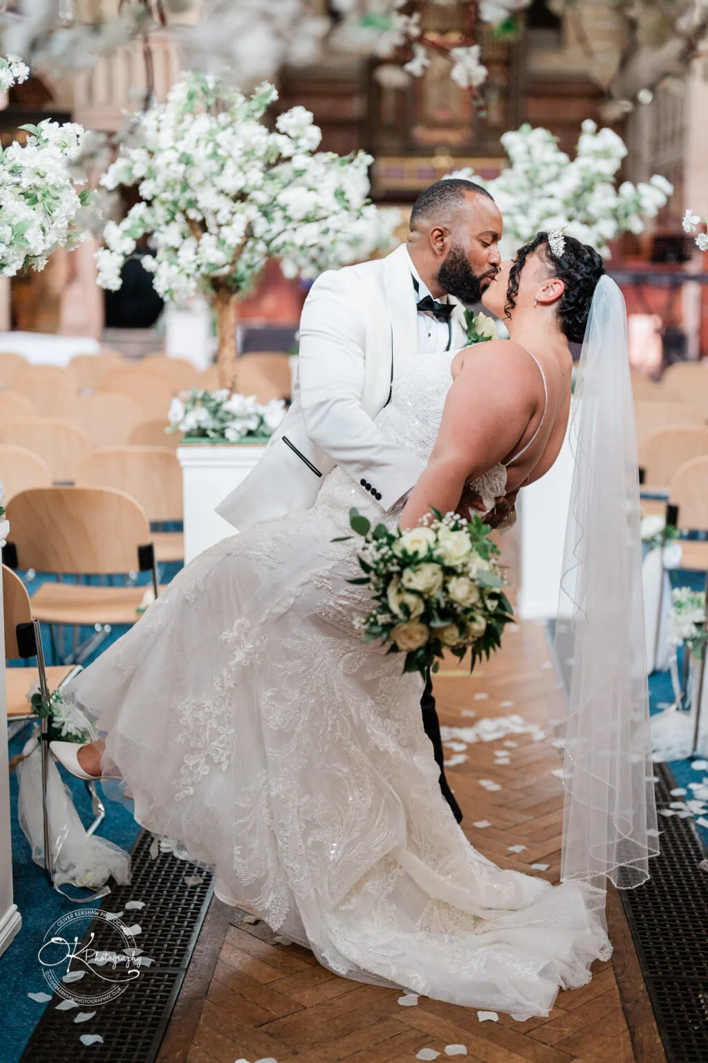 A groom in a white tuxedo dips and kisses his bride, who is wearing a white wedding gown and veil, inside a church decorated with white flowers.