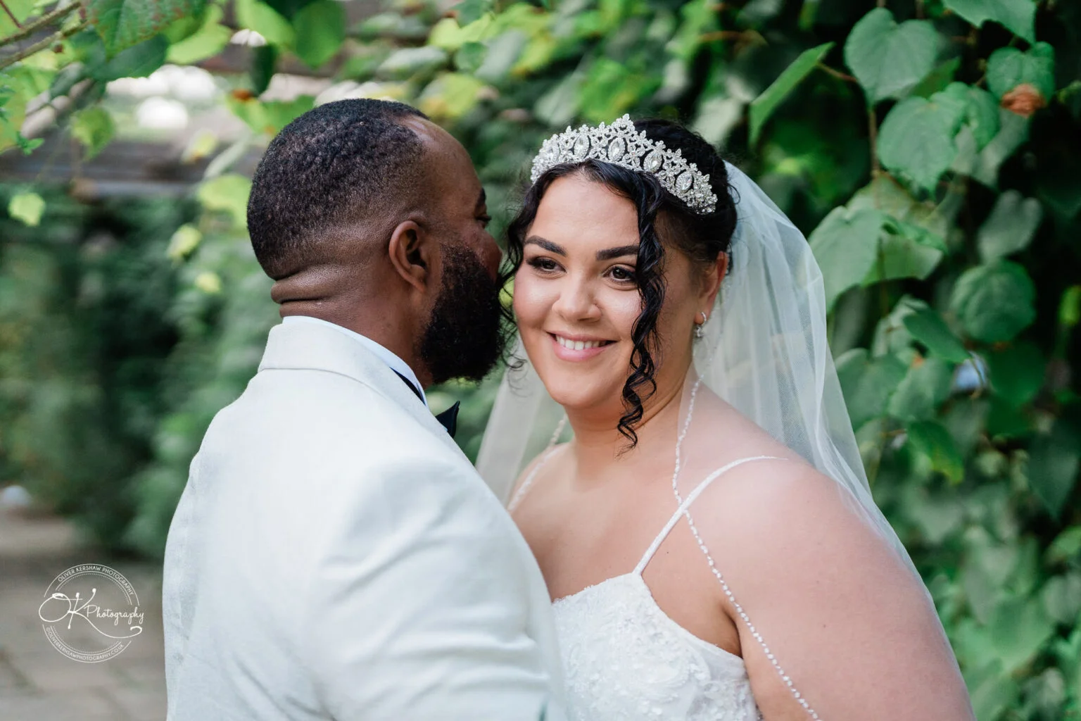 Bride and groom embrace on their wedding day, with the groom in a white suit and the bride in a white dress and tiara, surrounded by greenery.