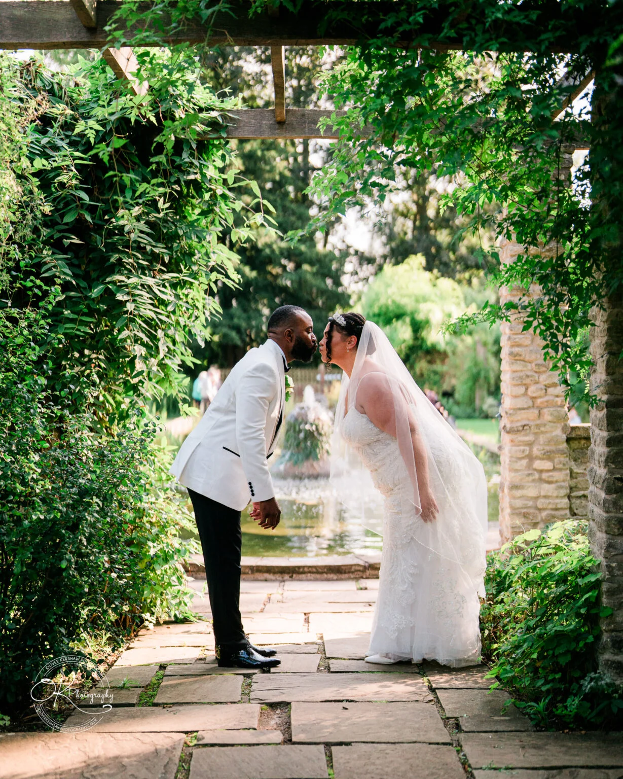 A bride and groom leaning in for a kiss under a green, leafy pergola, with a stone pathway and a scenic fountain in the background.