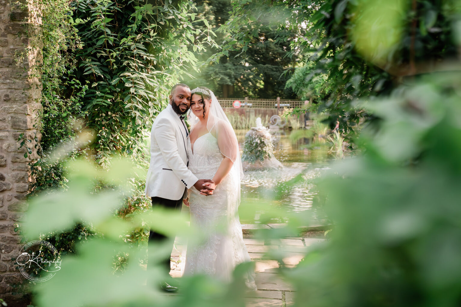 Symphony Rooms Wedding Photography Bride and groom holding hands in a lush garden setting with a fountain in the background, surrounded by greenery and stone structures.