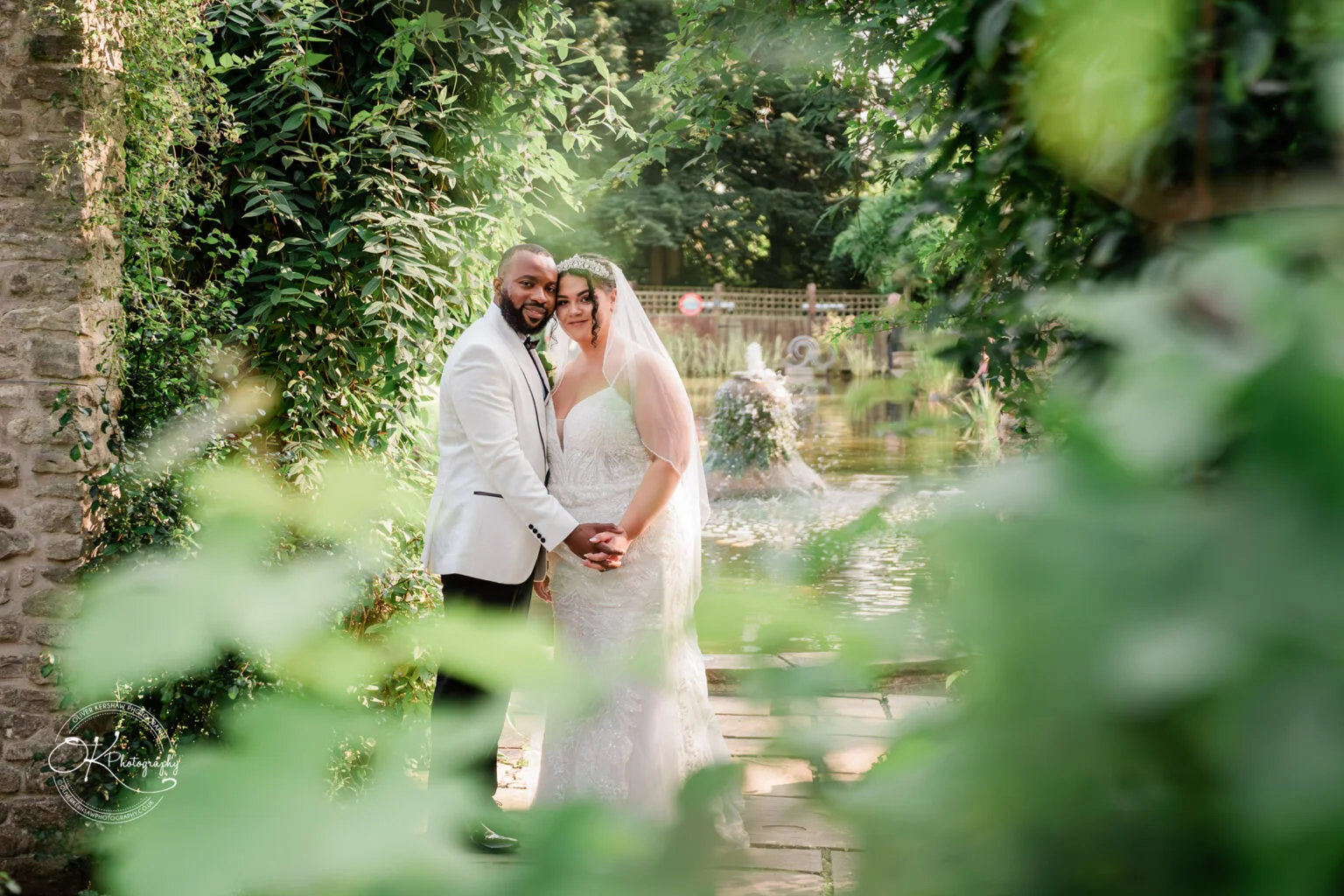 Bride and groom holding hands in a lush garden setting with a fountain in the background, surrounded by greenery and stone structures.