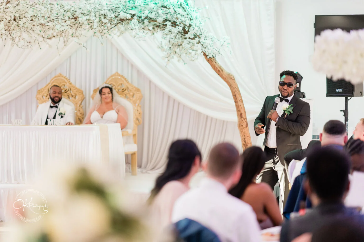 A man giving a speech at a wedding reception, with the bride and groom seated at a decorated table in the background.