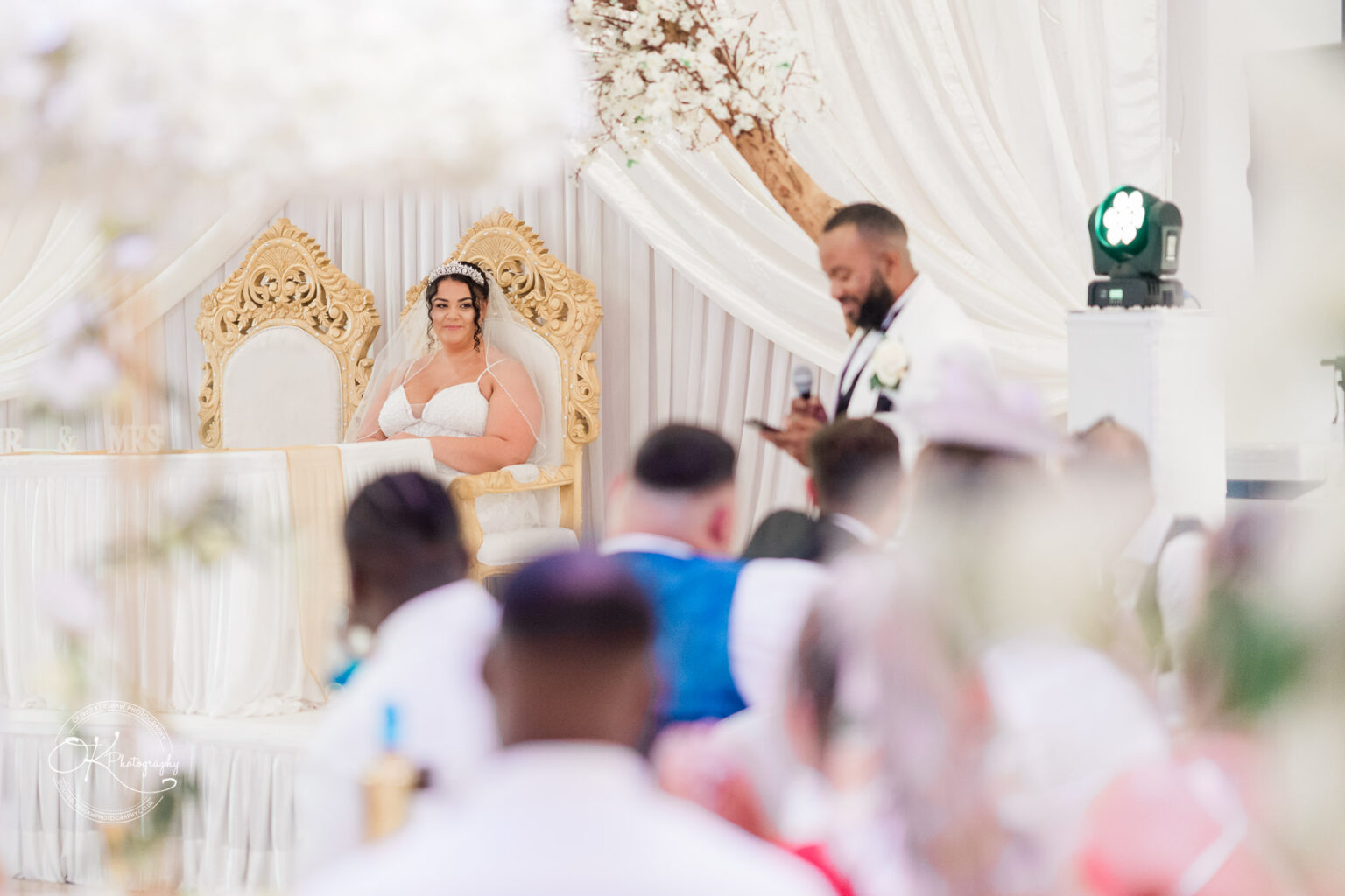 Symphony Rooms Wedding Photography Bride sitting on a decorative golden chair while the groom speaks into a microphone at their wedding reception surrounded by guests.