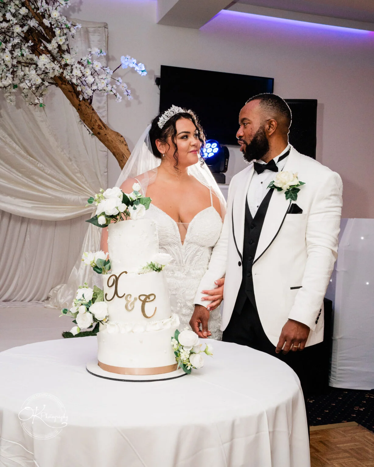 A bride and groom in wedding attire standing next to a tiered wedding cake decorated with white flowers.