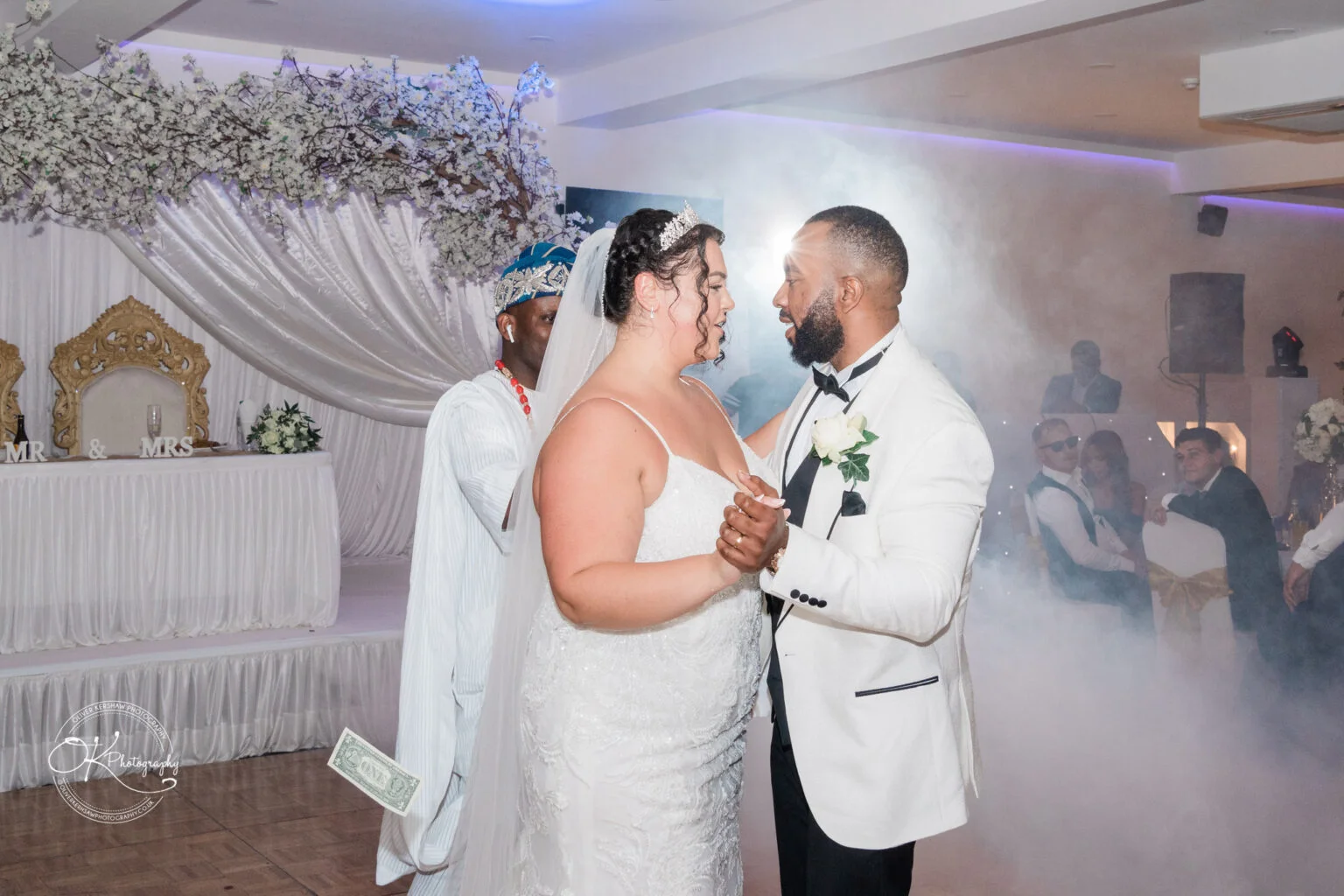 Bride and groom dancing at a wedding reception with a misty background, dressed in a white lace gown and white suit with bow tie and boutonniere, accompanied by a guest in traditional attire.