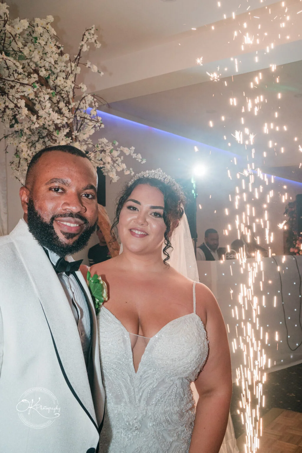 Newlywed couple smiling, with the groom in a white tuxedo and the bride in a white lace dress and tiara, standing in front of a sparking fountain.