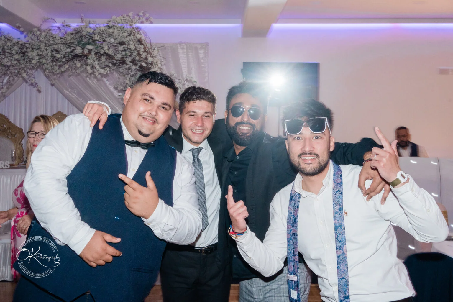 Four men in formal attire posing together in a brightly lit indoor setting, with two wearing sunglasses and one pointing.
