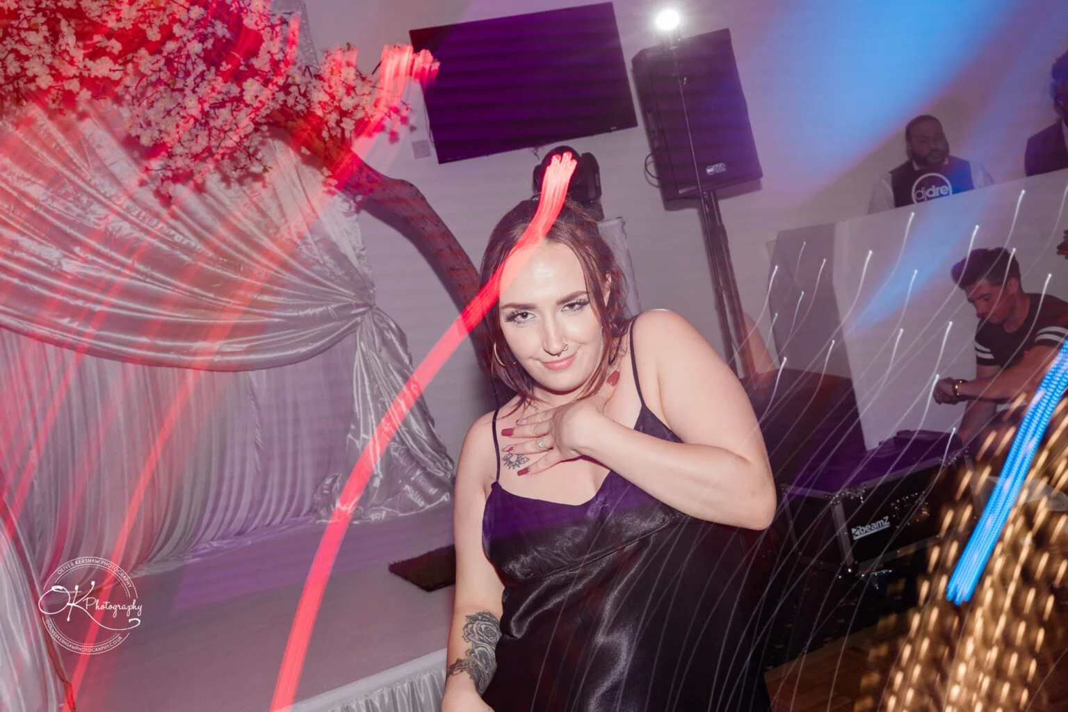 Person dancing at a party with colourful light trails, floral decorations, and a DJ in the background.