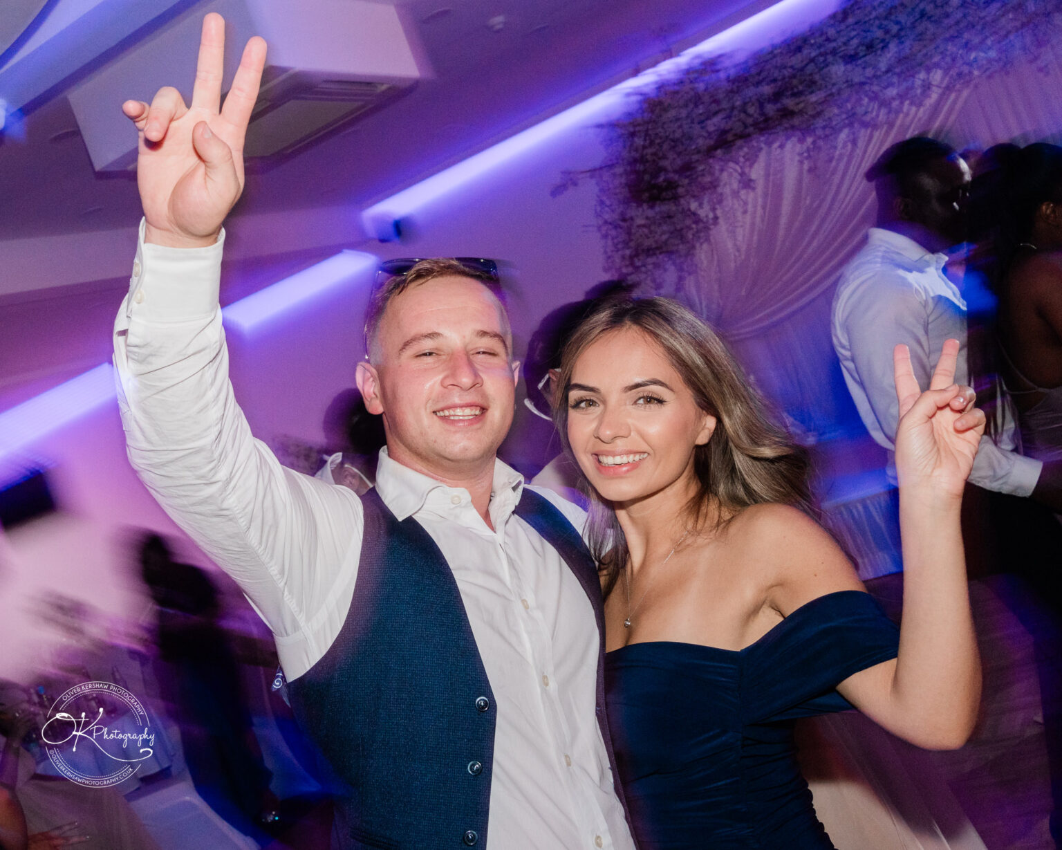 Symphony Rooms Wedding Photography A man and a woman smiling and posing with peace signs at a party, with blurred purple lighting in the background.