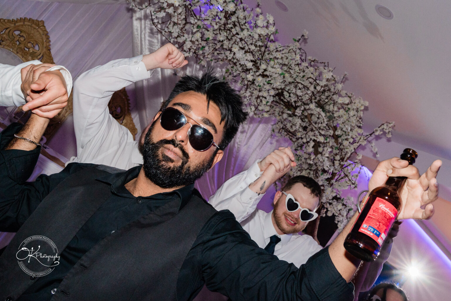 Symphony Rooms Wedding Photography A man with a beard and sunglasses, holding a beer bottle, poses with friends at a party.