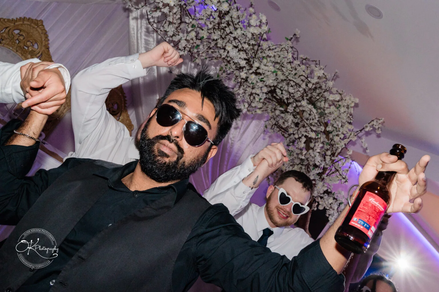 A man with a beard and sunglasses, holding a beer bottle, poses with friends at a party.