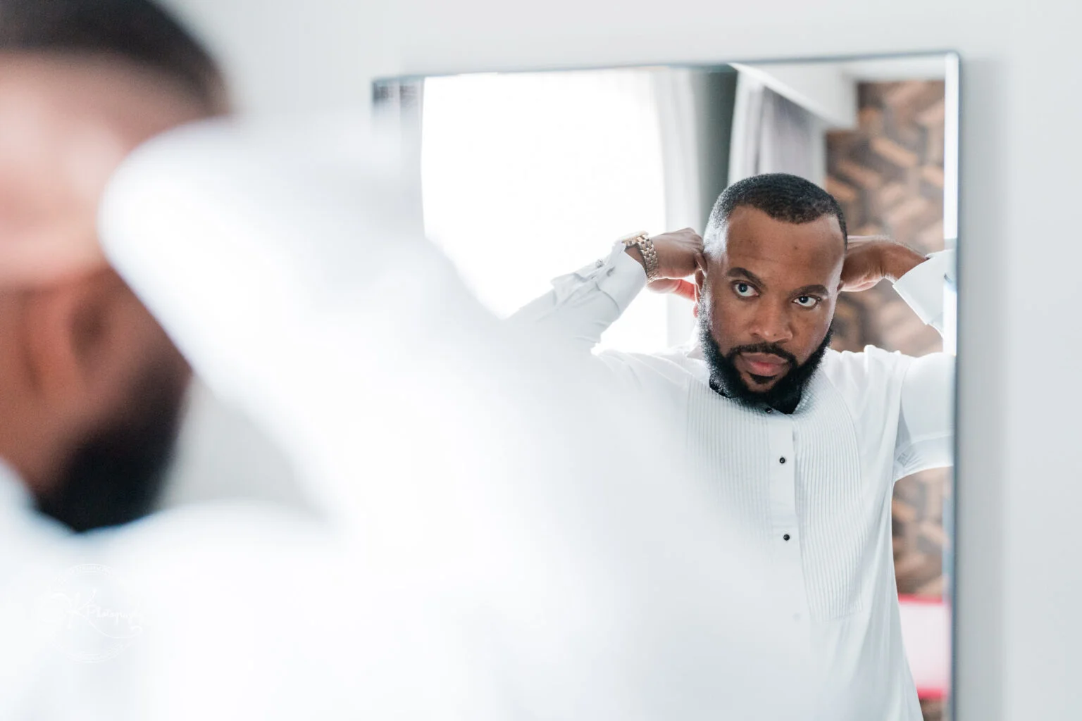 A man adjusting his collar in front of a mirror, wearing a white shirt and a wristwatch.