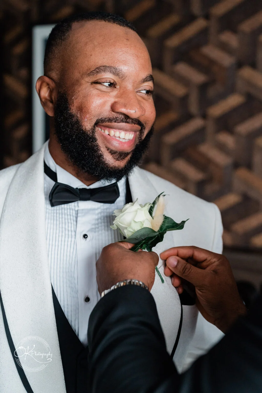 Smiling groom in a white tuxedo jacket with a black bow tie, having a white flower boutonniere pinned to his lapel.