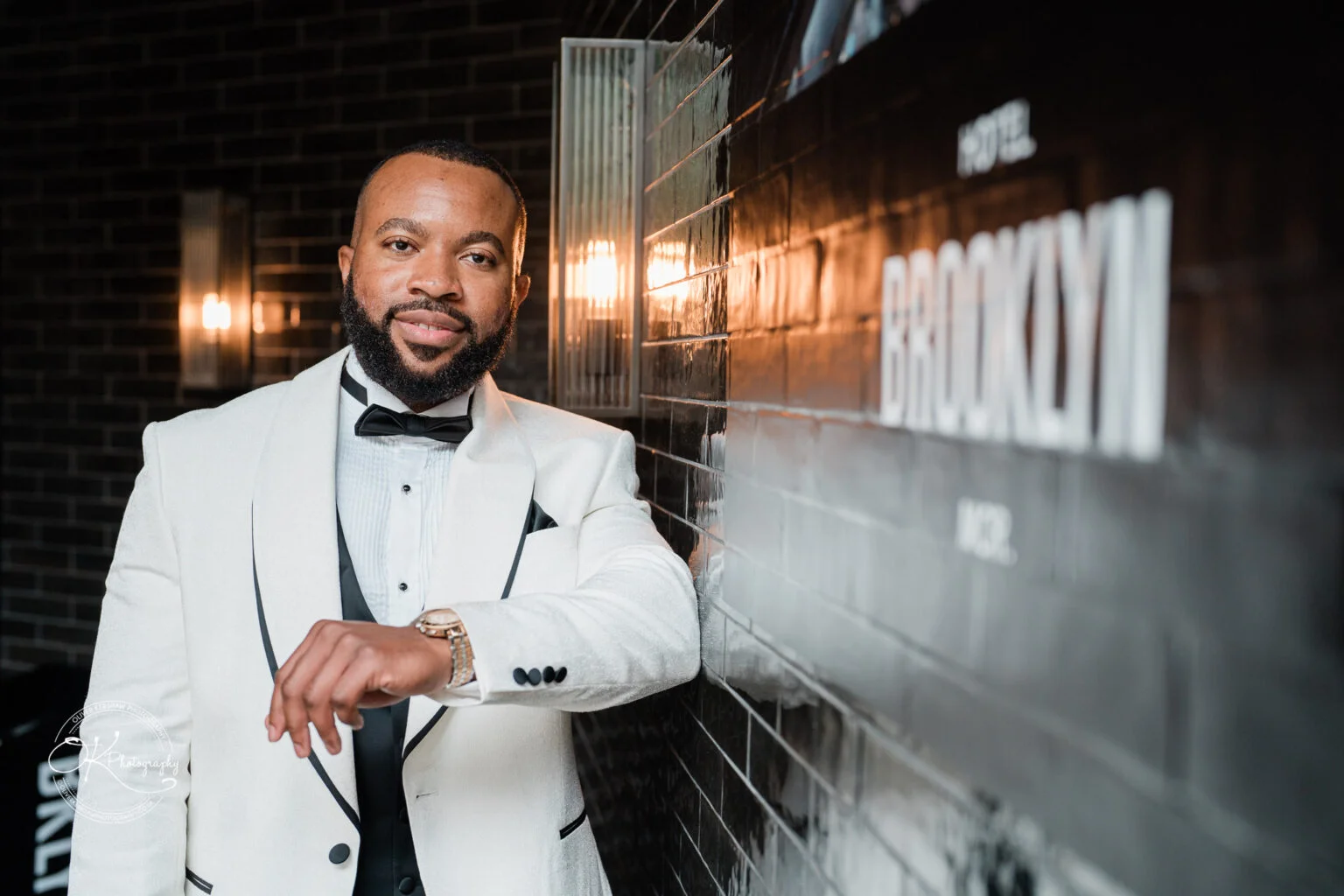 Man wearing a white tuxedo standing next to a wall lit by warm light fixtures, with the word "Brooklyn" visible in the background.
