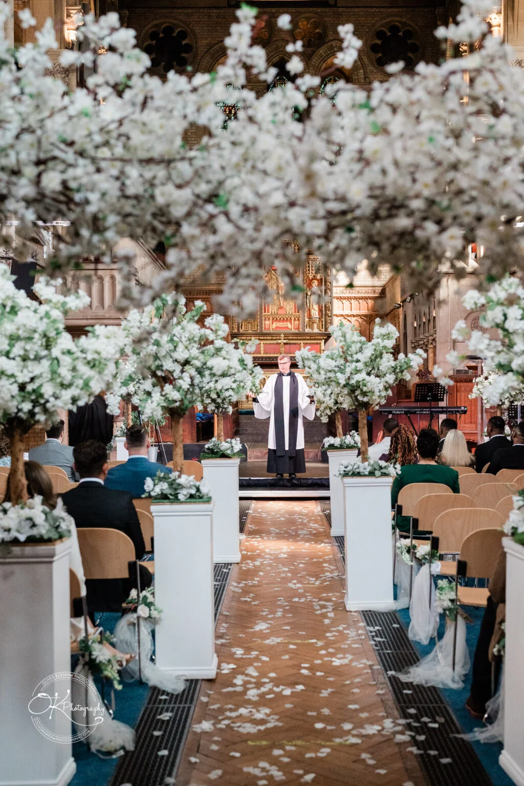 A wedding ceremony taking place in a church, with a priest standing at the altar. The aisle is adorned with flower arrangements and covered in white petals, and guests are seated on either side.