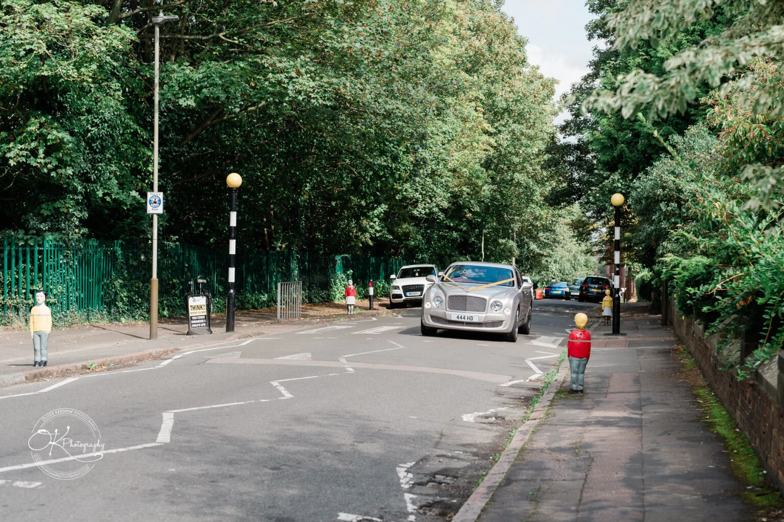 A silver luxury car driving down a tree-lined suburban street, with bollards shaped like toy soldiers on the sidewalk.