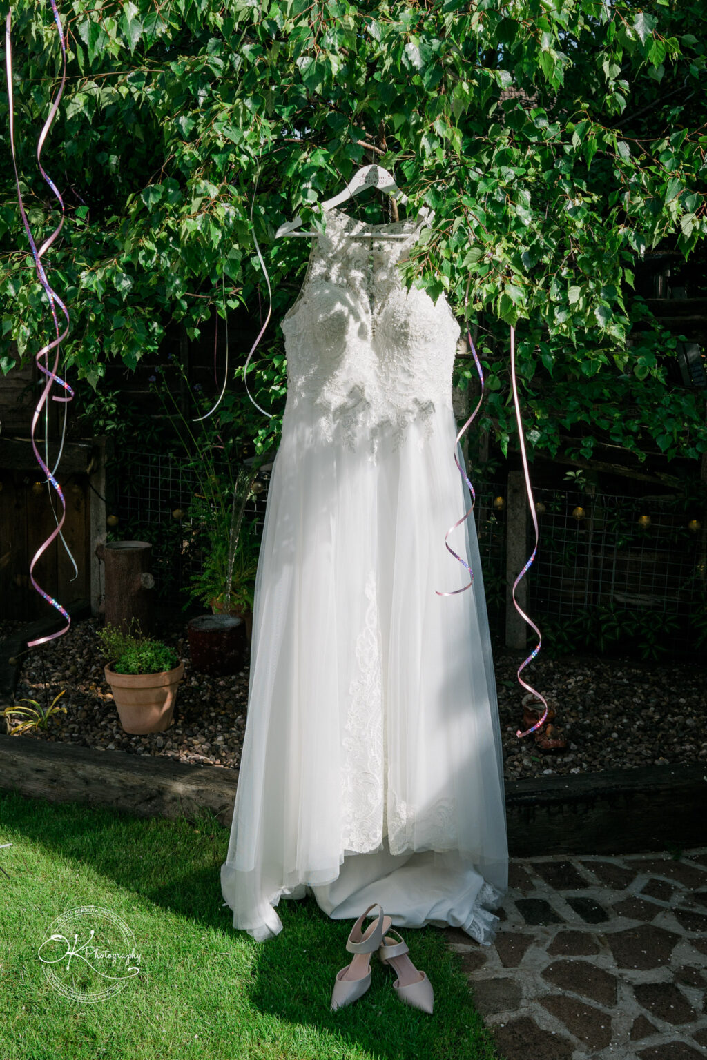 A white lace wedding dress hanging on a tree branch, adorned with streaming ribbon decorations. Pink shoes are placed on the grassy ground below.