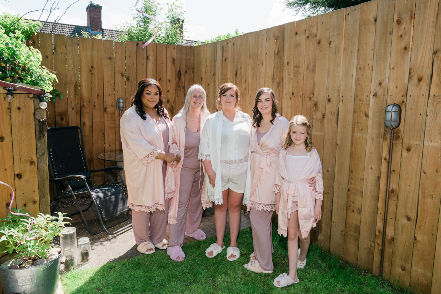 The Old Stables Wedding Photography A group of five women and girls wearing pink robes and slippers, standing together in a garden with a wooden fence in the background.