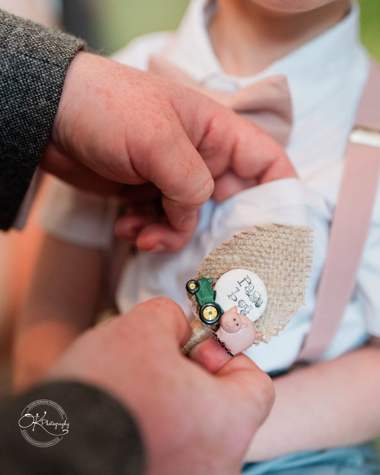 The Old Stables Wedding Photography A person pinning a rustic badge with miniature tractor and pig designs, labelled "Page Boy," onto a child's shirt with pink bow tie and suspenders.
