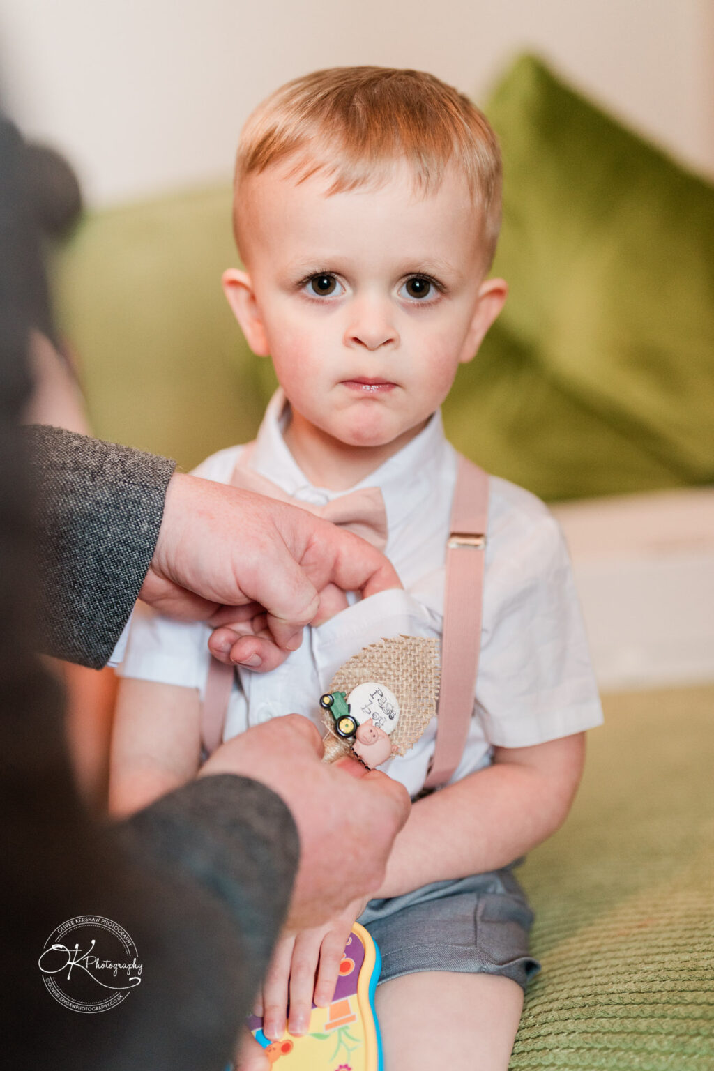 The Old Stables Wedding Photography A young boy dressed in a white shirt, bow tie, shorts, and suspenders, having a boutonnière pinned on his shirt by an adult.