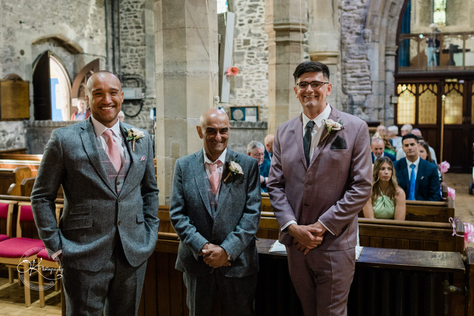 The Old Stables Wedding Photography Three men wearing suits and boutonnieres standing in a church with attendees seated in the background.