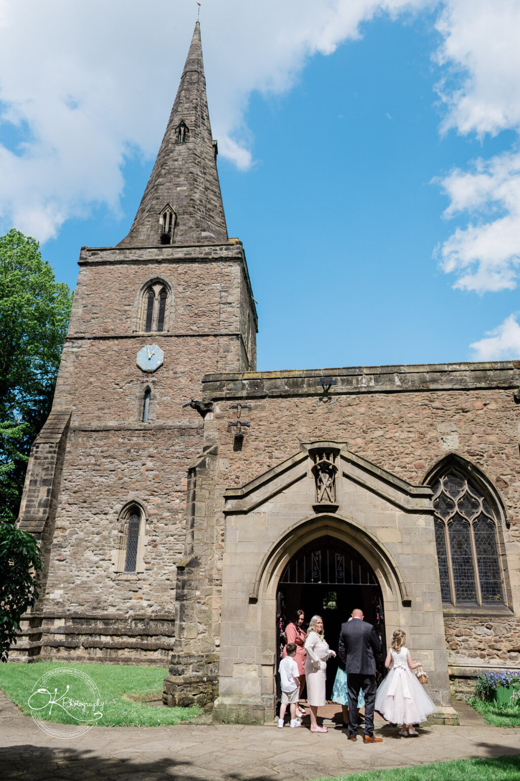 The Old Stables Wedding Photography Guests standing outside the entrance of a stone church with a tall spire on a sunny day.