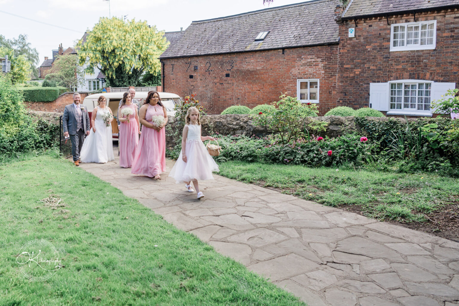 The Old Stables Wedding Photography A bride, groom, bridesmaids, and a flower girl walking along a garden path next to a brick house.