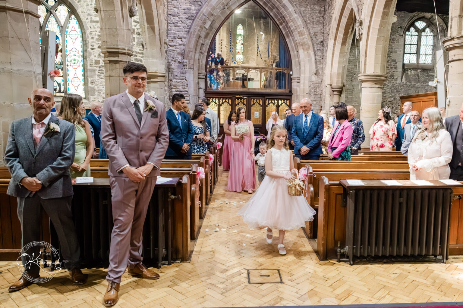 The Old Stables Wedding Photography Flower girl walking down the aisle in a church, followed by a bridesmaid holding flowers, with guests standing on either side and a groom waiting at the altar.