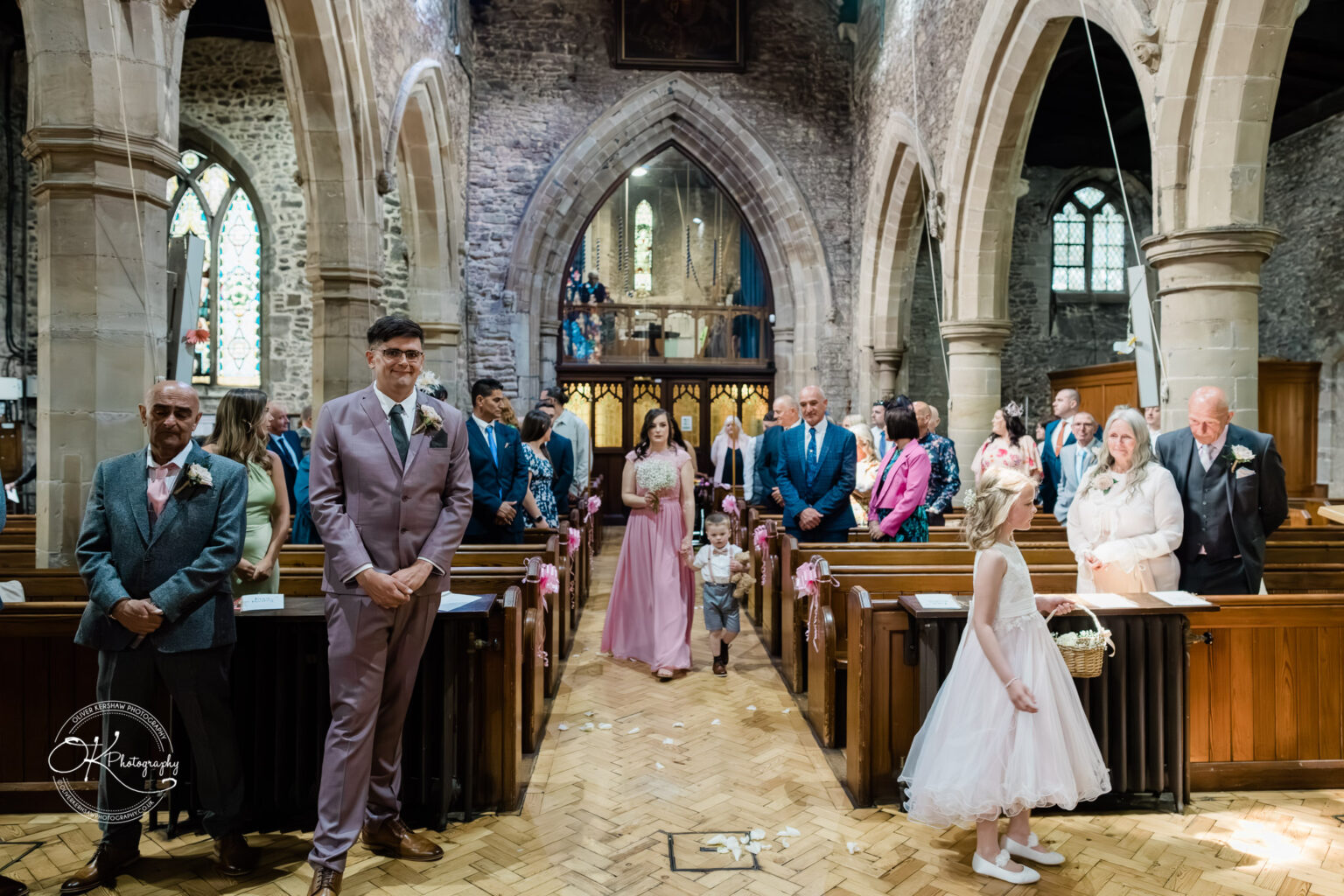 The Old Stables Wedding Photography A wedding ceremony in a church with guests seated in pews, the groom standing at the front, and the bridal party, including a flower girl and a young boy, walking down the aisle.