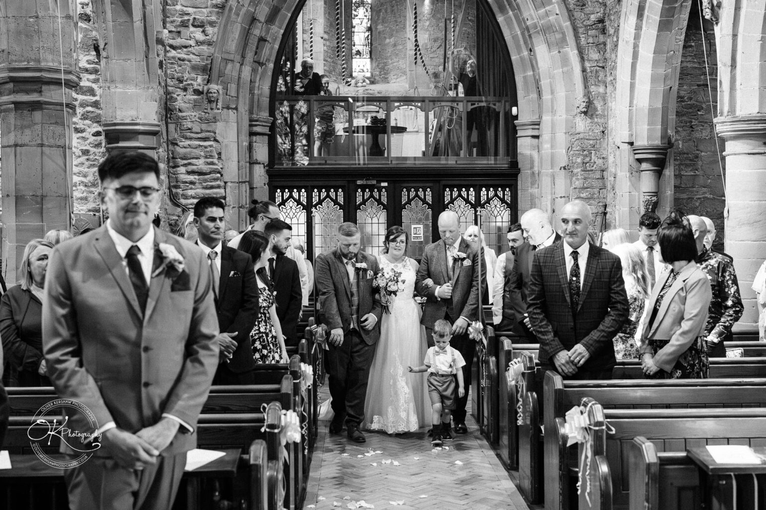 The Old Stables Wedding Photography A bride walks down the aisle in a church, accompanied by two men, with guests looking on and a groom waiting at the altar.