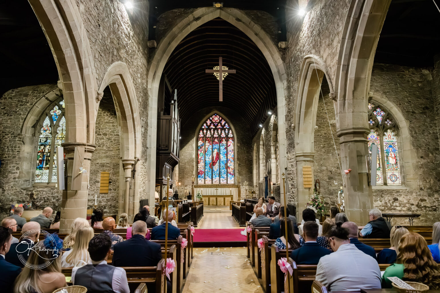 The Old Stables Wedding Photography Interior of a stone church with stained glass windows and wooden pews, filled with guests seated and facing the altar during a wedding ceremony.