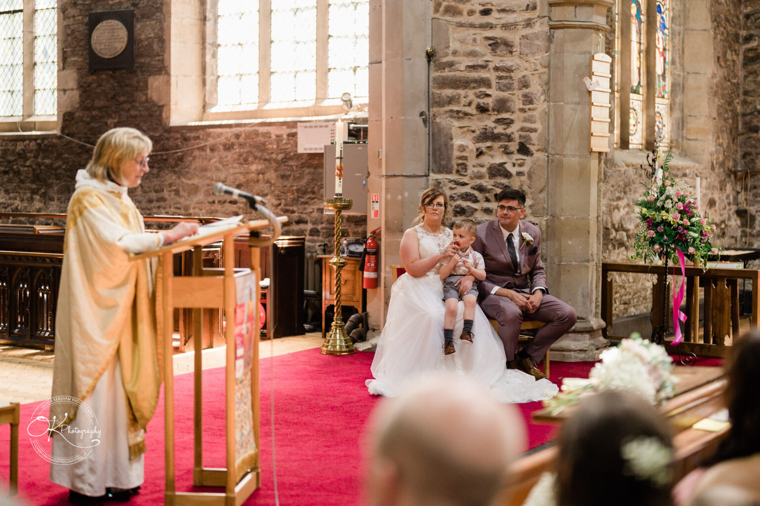 The Old Stables Wedding Photography A priest addresses a congregation in a church as a bride, groom, and young child sit nearby during a wedding ceremony.