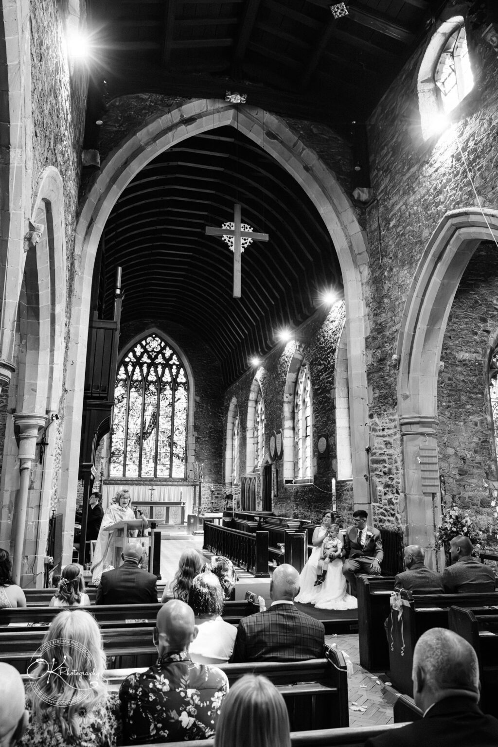 The Old Stables Wedding Photography Black and white wedding photograph inside a historical stone church with vaulted ceilings, where the couple and guests are seated, and a woman is speaking at the lectern.
