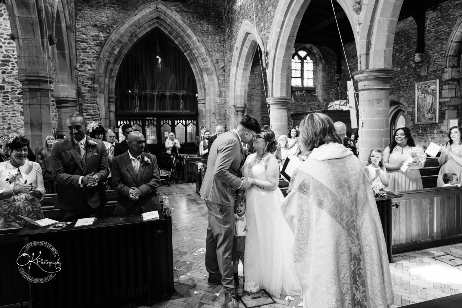 The Old Stables Wedding Photography A bride and groom share a kiss during their wedding ceremony in a historic stone church, surrounded by smiling guests and officiated by a robed minister.