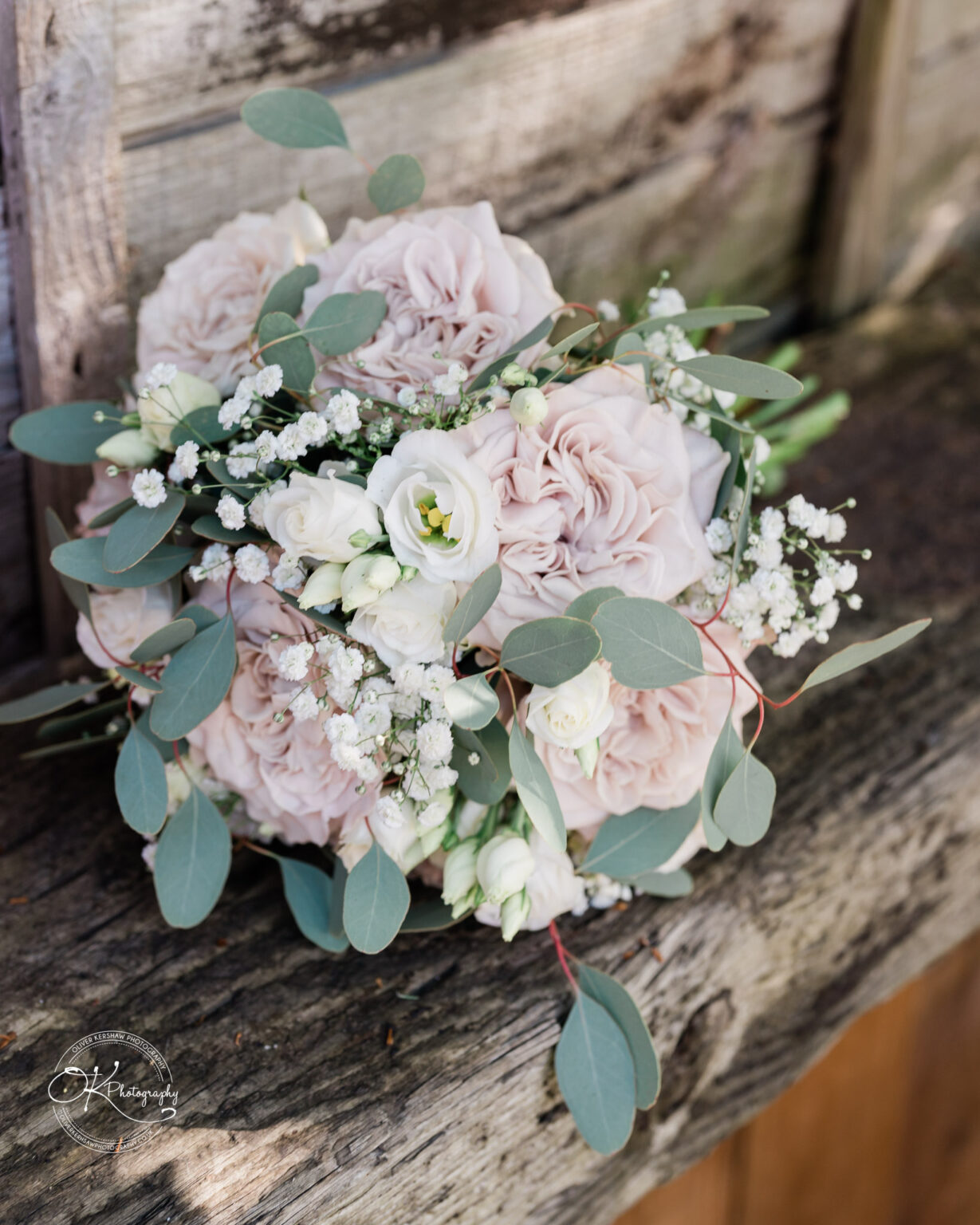 The Old Stables Wedding Photography A bouquet of light pink and white roses with eucalyptus leaves and baby's breath flowers, resting on a rustic wooden surface.