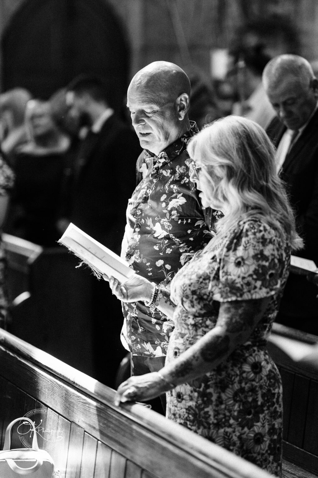 The Old Stables Wedding Photography A candid black and white photograph of a man and woman standing in a church pew, reading from a book during a wedding.