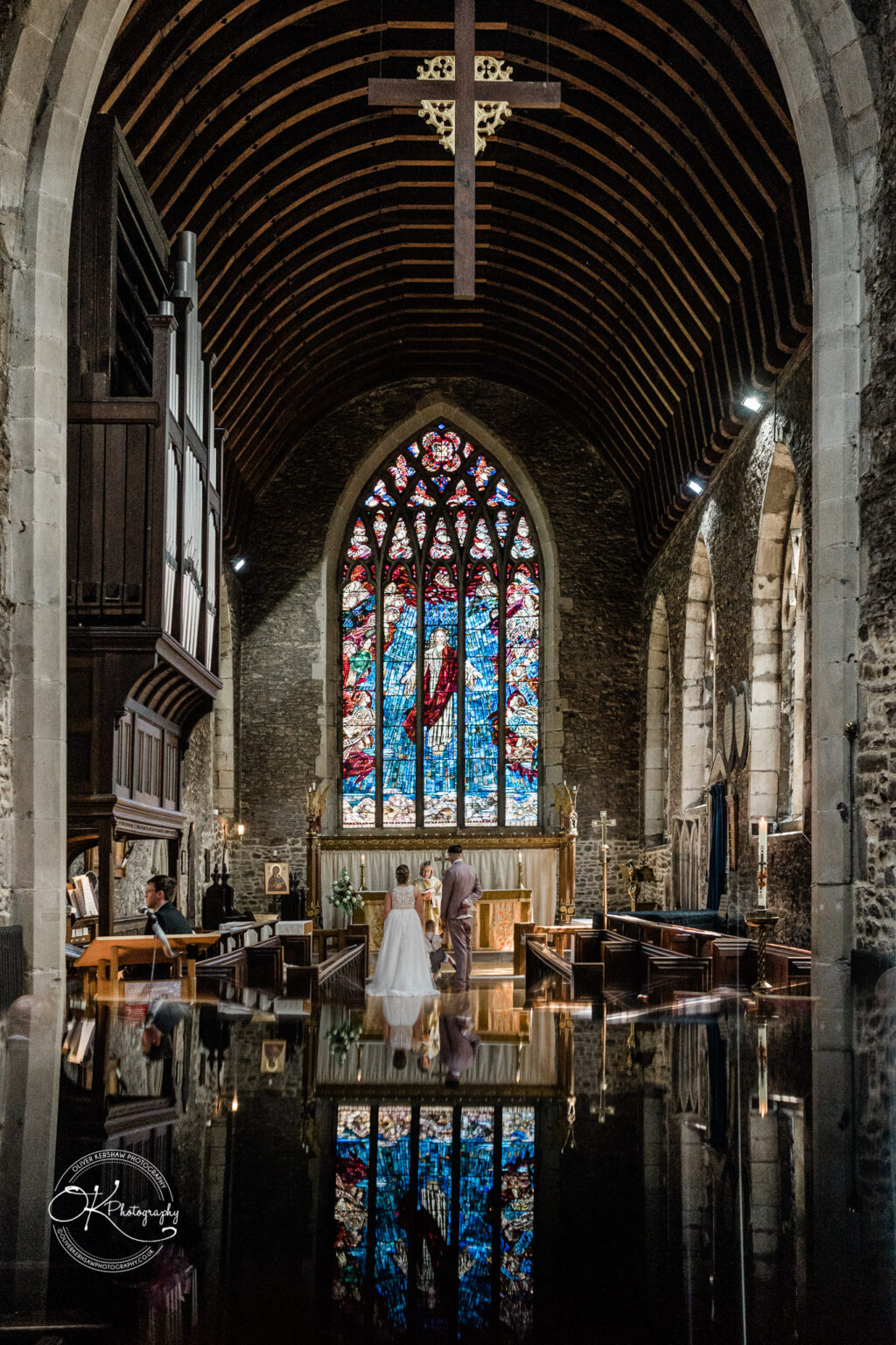 The Old Stables Wedding Photography Interior of a church featuring a wedding couple standing before the altar, with a large stained glass window and a cross overhead.