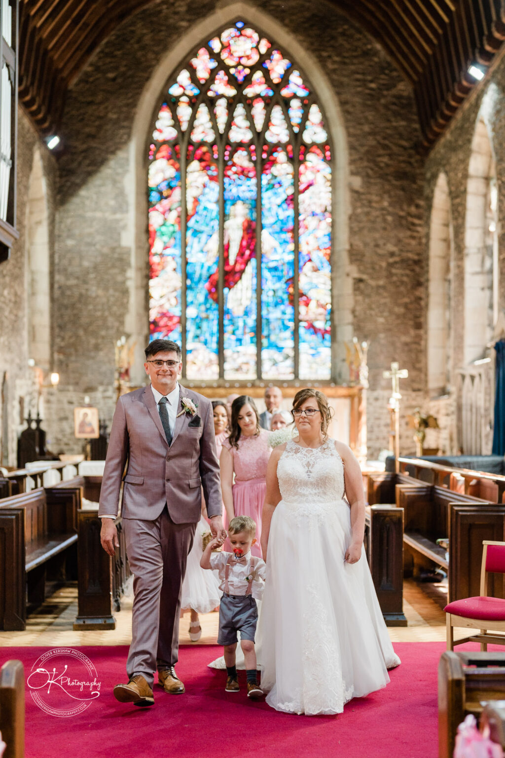 The Old Stables Wedding Photography Bride and groom walking down the aisle with a young child after the ceremony in a church with a large stained glass window in the background.