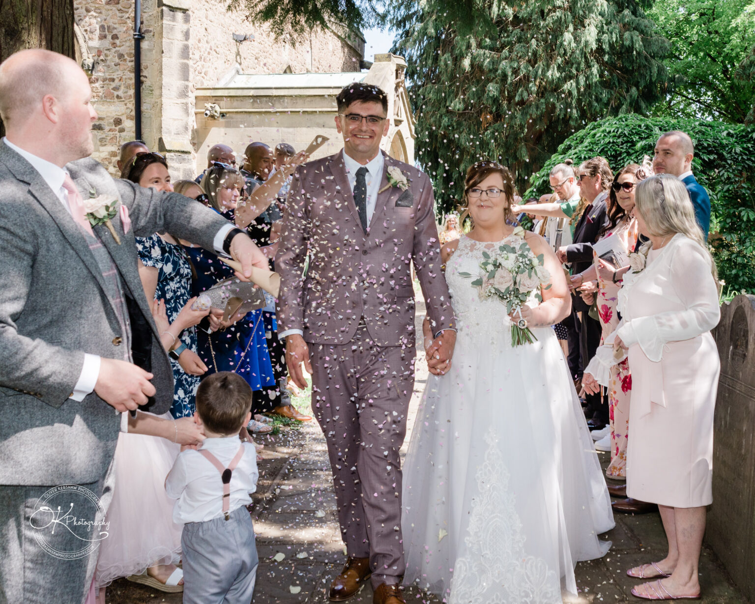 The Old Stables Wedding Photography Bride and groom walking down an aisle outdoors, surrounded by wedding guests throwing confetti.
